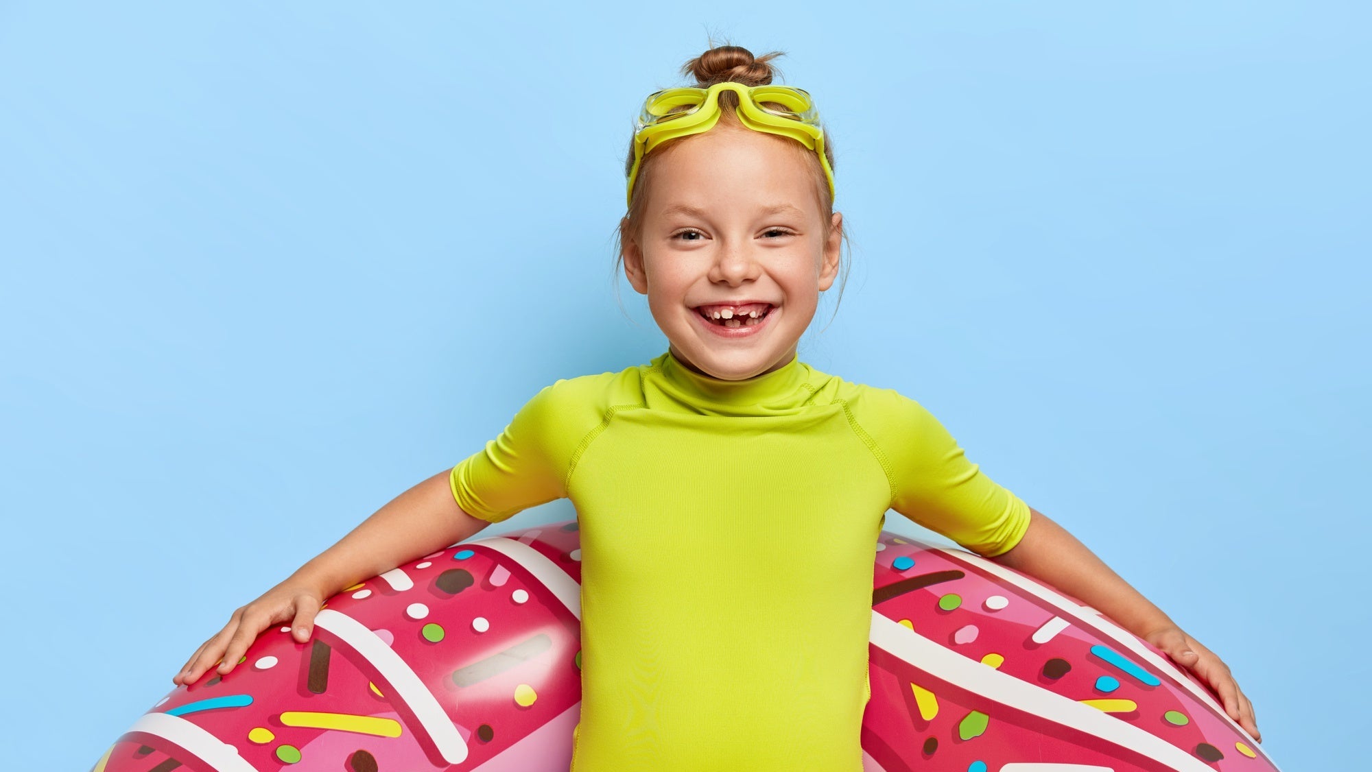 Child in green shirt and blue shorts with a pink inflatable donut float against a light blue background