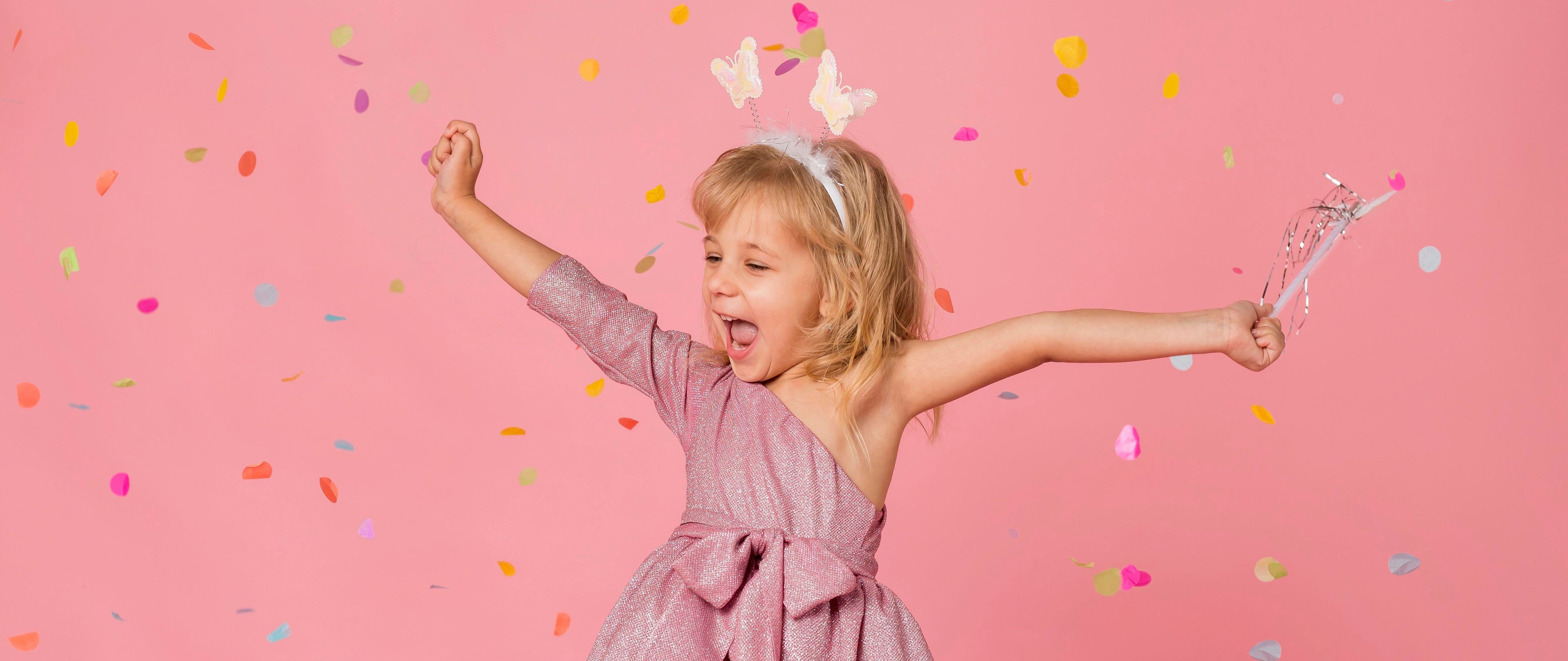 Young girl in a pink dress with a wand and butterfly headband against a pink background with confetti.
